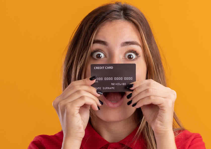 Imagem mostra uma mulher sobre um fundo amarelo segurando um cartão de crédito em frente a seu rosto com os olhos arregalados.