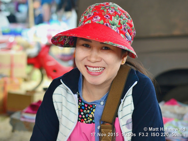 Matt Hahnewald; Facing the World; Asia; Northern Thailand; Chiang Dao; Tuesday market; people; closeup; portrait; street portrait; headshot; smiling; Thai woman; Thai smile; market woman; friendliness; Land of Smiles; world cultures; travel; tourism; eye contact; Nikon DSLR D3100; 50 mm prime lens; colour; face; market; hat; outdoor