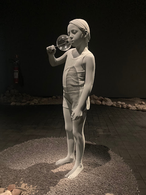 A lifelike grey sculpture of a young girl blowing a bubble.