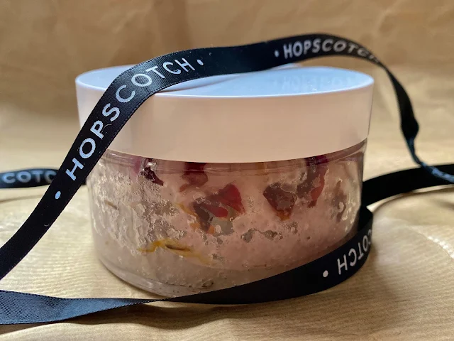 A glass jar with bath salts containing rose petals with a black ribbon saying HOPSCOTCH on it