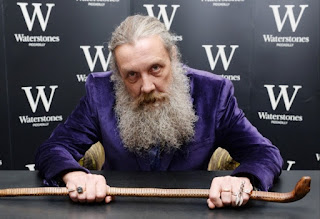 Alan Moore seated at a book store signing table with Waterstones logos forming the backdrop. Alan is wearing a purple velvet jacket and holding a magician's staff, horizontally, towards the camera
