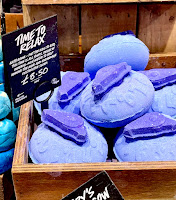 A photo of a light square brown box containing some blue spherical bath bombs with dark blue handles with a black rectangular card that says time to relax bath bomb lush in white font on a bright background