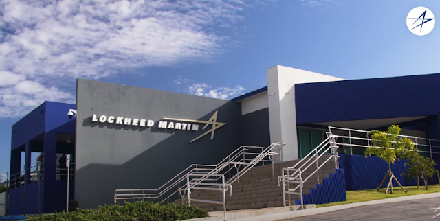 Lockheed Martin building with sleek modern design, featuring blue and gray exterior and prominent logo. Bright day with scattered clouds.