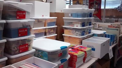 Stacked clear bins with white lids, containing miscellaneous items, along with books in stacks or upright inside lidless boxes covering a large table-top surface