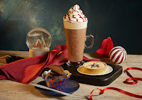 A wide angled photo of a tall cylindrical glass filled with dark brown hot chocolate, white whipped cream and red sauce next to a small white circular plate with a light brown mince pie with a star on top of it next to a dark brown chocolate muffin on a dark brown rectangular table on a bright background