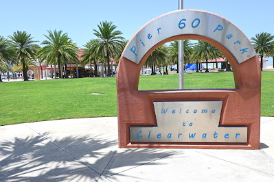 A sign for Clearwater Beach's Pier 60 Park. It is a brownish color, is in the shape of an upside down "U," and has a square bottom. In an arch across the top, and in blue pain with a silver background, it reads: "Pier 60 Park." On the bottom of the sign, it reads: "Welcome To Clearwater"