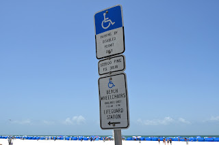 A disabled parking sign with the beach and blue sky behind it. It has three sections, which read, "Parking by disabled permit only. $255.00 fine. F.S. 318.18. Beach wheelchairs available daily. 9:30 AM - 4 PM. Lifeguard Station.