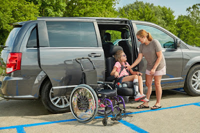 A woman helps her daughter transfer out of their dark gray minivan. Her daughter is seat belted into a transfer seat, which has extended out of the vehicle and closer to the ground. The mother reaches to unfasten the seatbelt. She is wearing a gray top, an off-white skirt, black sandals, sunglasses, and has her blonde hair in a ponytail. Her daughter is wearing a pink shirt, blue shorts, white sneakers, sunglasses, and braces around her shins. They are looking at each other smiling. A purple, manual wheelchair is situated next to the little girl.