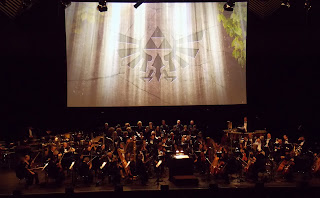 photo of the stage during rehearsal, there is a large screen behind the orchestra with a Hylian Crest displayed