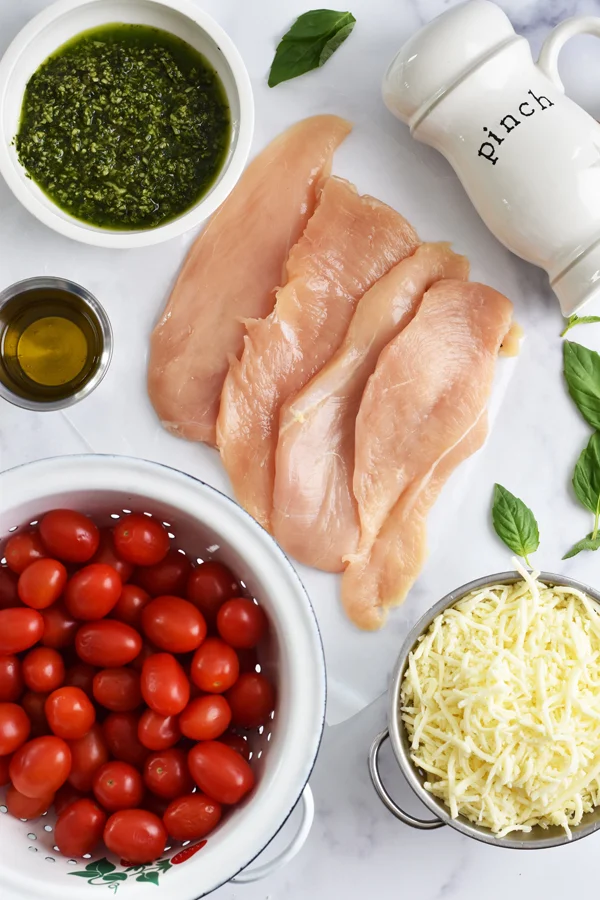 A colorful assortment of ingredients for making baked pesto chicken, including chicken cutlets, homemade pesto sauce, grape tomatoes, mozzarella cheese, fresh basil leaves, olive oil, balsamic glaze, and kosher salt.