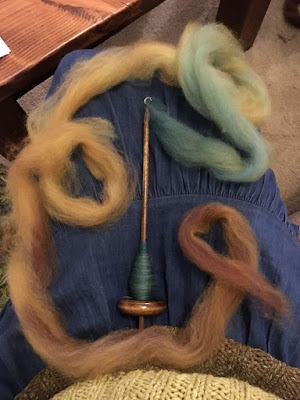 A photo from above of the photographer's lap, showing a gathered denim skirt and the edge of a color-blocked olive-and-apple-green sweater. A wooden bottom-whorl spindle rests in the center of the photographer's lap, with the whorl toward their body, and a loose rope of wool coiled around it, teal-blue at the hook of the spindle through gold and into cinnamon and russet at the trailing end.