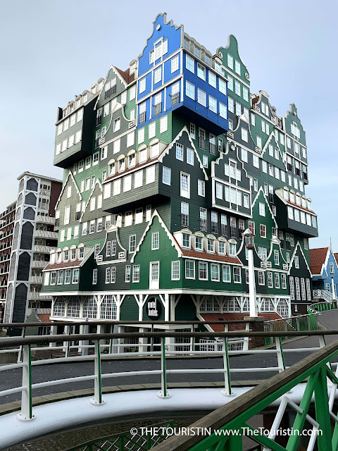 11-storey building that looks like a pile of several green wooden houses with white windows and red roofs plus one prominently placed blue house at its corner on the top.