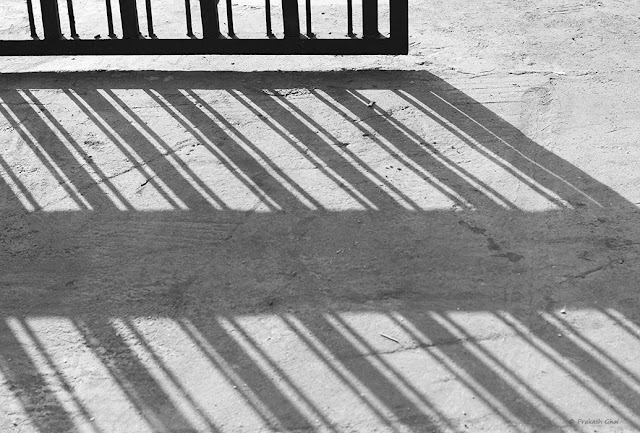 A Black and White Minimalist Photo of the Long Shadow of a Metal Gate being cast on the Street.