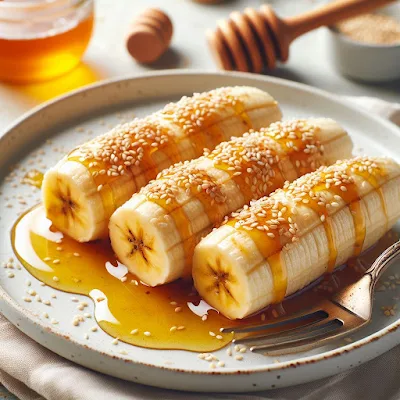Auf dem Bild sieht man einen Teller mit vier Bananen die gebraten sind. Sie sind mit Honig beträufelt und mit Sesam bestreut. Der Nachtisch sieht sehr lecker und appetitlich aus.