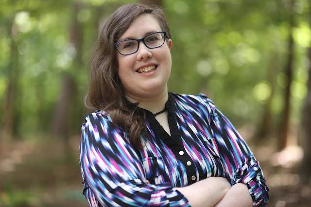 Photo of the author, a white woman with brunette hair, wearing bold blue glasses and a multicolor shirt, standing in a forest.