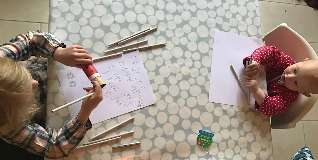 A 5 year old girl sitting at the table opposite her sister at the dining table. They are both drawing with colouring pencils