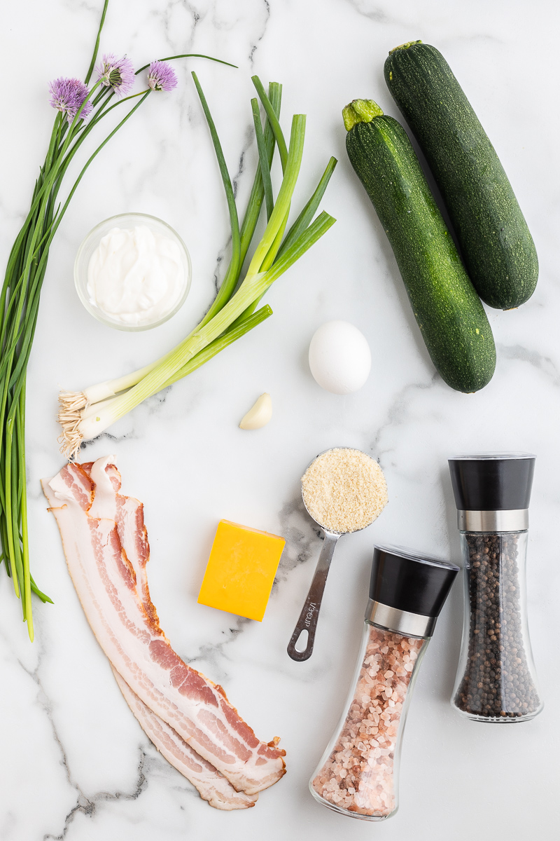 Overhead photo of the ingredients needed to make Keto Cheesy Zucchini Fritters with Bacon on a white marble counter.