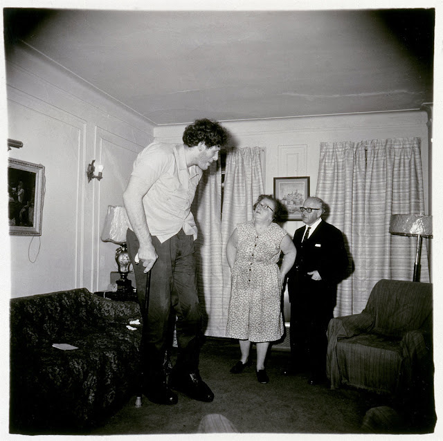 Foto em preto e branco, dentro de uma casa, na sala, com um casal idoso, olhando para um homem gigante