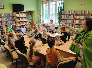 Tło sala biblioteczna: regały z książkami, regał z kolorowymi czasopismami. Grupa dzieci siedzących przy stolikach. Pani bibliotekarka stoi przed grupą dzieci i wyznacza osobę do odpowiedzi. dzieci podnoszą ręce do góry w celu odpowiedzi na pytanie zadane przez panią bibliotekarkę.
