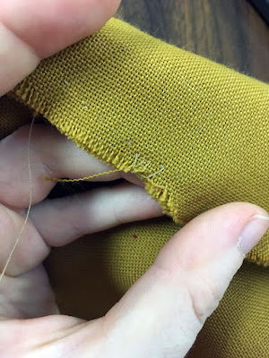 A close-up of a slightly-frayed seam in mustard-brown fabric, held up by a white hand and stitched with tan thread.