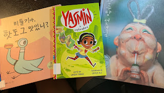 Photo of The Pigeon Finds a Hot Dog translated into Korean, Yasmin la exploradora, and a Korean storybook with an older lady on the cover sipping a drink through a straw. You can only see her from the shoulders up, but she is nude since the story takes place in a bath house or spa.