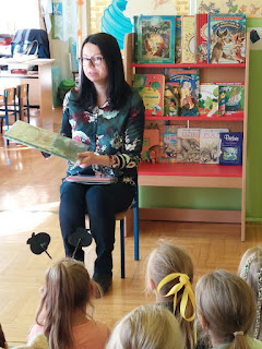 Pani bibliotekarka siedzi na krześle i pokazuje dzieciom książkę. tło: regał z książkami.