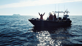 Kolme ihmistä veneessä, yhdellä kädet ilmassa.