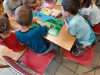 Na zdjęciu grupa dzieci oglądająca książki. Książki ułożone są na drewnianym stoliku, przy którym siedzą dzieci.