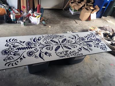 A white plywood panel with a bold folk art floral and bird pattern in deep indigo-purple, propped on two grey plastic totes in a cluttered garage.