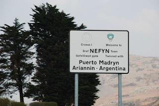 A black and white road sign gives place names in Spanish and Welsh. Trees and a mountainside are glimpsed behind it.