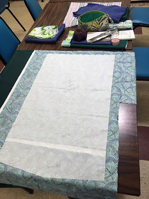 The backside of a turquoise and navy art deco print fabric, laid out on a table over a dark hunter green fabric, with a large, rectangular tissue paper pattern piece pinned to it, and a variety of stacks of green and blue fabrics, red pincushion, silver shears, and other sewing apparatus in the background.