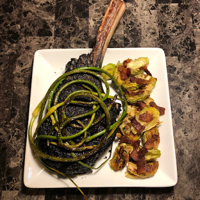 Prime tomahawk ribeye grilled Pittsburgh rare, topped with grilled garlic scapes along with bacon roasted Brussels sprouts