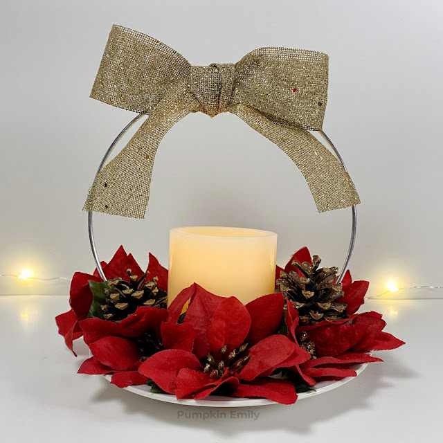 A hoop centerpiece for Christmas with red flowers, gold bow, pinecones, and a LED candle.