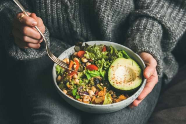 Healthy vegetarian dinner. Woman in jeans and warm sweater holding bowl with fresh salad, avocado, grains, beans, roasted vegetables