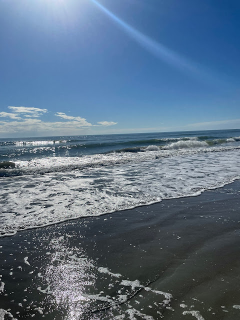 A picture of the water. There are bubbles all around us as the waves finish there ascent up the shoreline. The sun is beaming down on the water. There are deep ripples in the sand from where the waves have pushed themselves back and forth over the land.