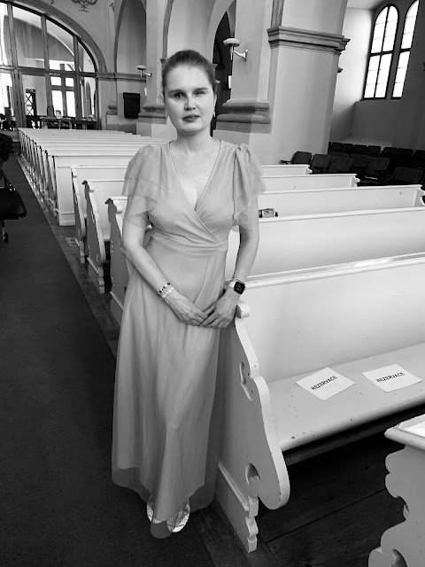 Linda stojí v kostele, za ní je řada lavic. Fotka je černobílá