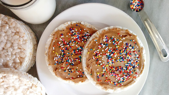 a close up of two cookie butter and sprinkles rice cakes sitting on a plate next to a stack of rice cakes, a cup of milk, and a measuring spoon full of sprinkles