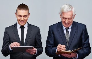 Young Man in Suit Holding Tablet Stands next to Old man in Suit With Pen and Paperr Holding Tablets