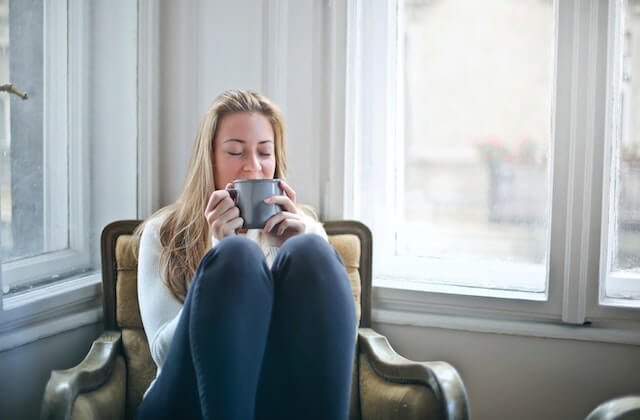 Blonde woman looking happy and content sitting on a chair by the window drinking coffee