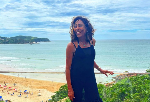 Mulher sorridente, apoiada em parapeito de vidro com o mar ao fundo