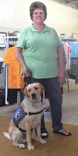 Picture of Toby wearing his coat in a sit-stay beside the consignment store owner (who is wearing jeans & a green shirt)