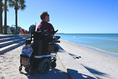 Kyle is shown in his white, Quickie S-636 power wheelchair. He's wearing a maroon- and navy-striped shirt, and is facing away from the camera. He's wearing sunglasses, and looking to the right, over the ocean. His wheelchair is positioned on a concrete path that runs along a stretch of the beach. To his left are a few palm trees, as well as a flight of concrete stairs and an aluminum hand rail.