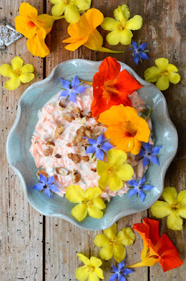 Cremiger Karottensalat mit Joghurt in einer grünblauen Schüssel mit gewelltem Rand. Rundherum und auf dem Salat liegen Blüten in den Farben gelb, orange und blau.