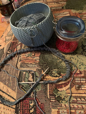 A circular knitting needle with sage green stitches casted onto it, sits in front of a blue-gray bowl holding the ball of sage colored yarn. The bowl looks as if it were knitted. The bowl is next to a sealer jar containing red glass seed beads. It has a transparent cobalt lid and silver hardware.