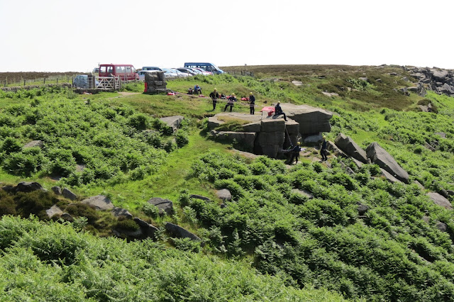 A large outcrop of gritstone in the bracken. A group of helmeted and roped up MRT members are practising hoisting someone up the rocks.