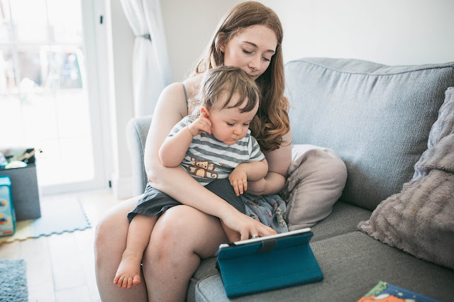 toddler boy sitting on his mother's knee. Mother is pointing to ipad screen and child is watching and touching his ear