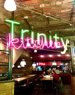 A bright yellow neon sign saying Trinity Kitchen with the kitchen in cursive white font on a bright background. 