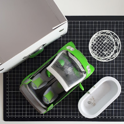 Flat lay of an empty storage box on a cutting mat with a one-twelfth scale modern miniature smart car, bath and round wire table on it.