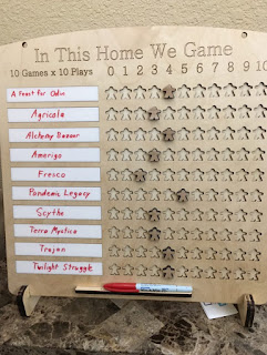 A wooden board, laser etched, with feet to allow it to stand up. Along the top it reads, 'In This Home We Game.' Beneath that it says '10 Games x 10 Plays,' next to the numbers zero through ten. The main portion of the board is ten strips of dry-erase areas on which are written the names of ten games. Beside these are rows of ten meeple-shaped cut-out spaces, with meeples placed in each row to indicate how many times that game has been played. There is a ledge at the bottom on which to place a dry-erase marker.