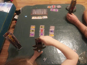 A game of Swordcrafters in progress, seen from above as two pairs of arms manipulate their swords. A third sword stands nearby on the table. Some tiles are still on the table waiting to be claimed, as the score tracker and three sword magic cards sit nearby.